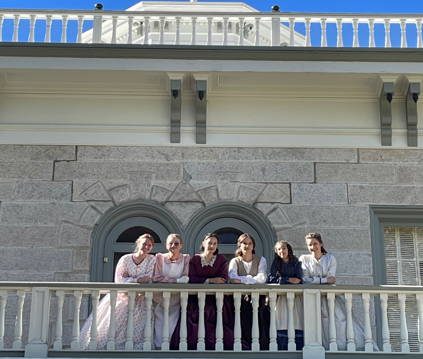 Six living history characters standing on the Bowers Mansion balcony wearing Victorian dresses.